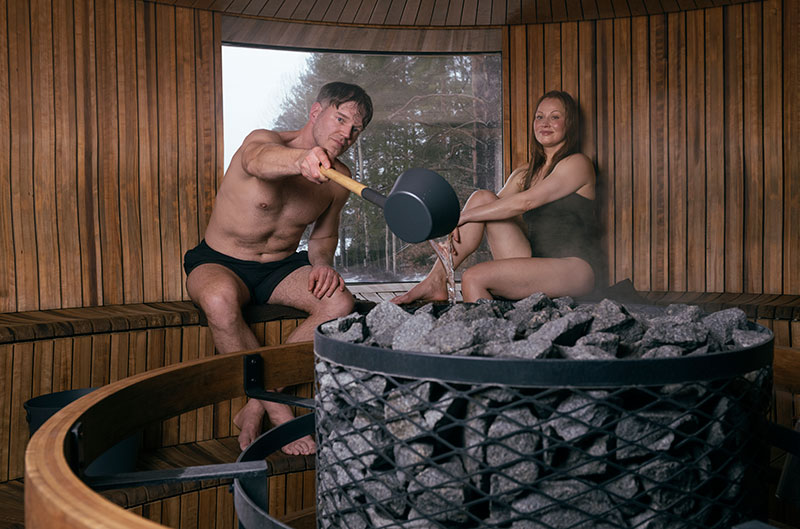 Een man giet met een pollepel water over de hete saunastenen, een vrouw zit ernaast; een stomende, ronde houten sauna met een raam.